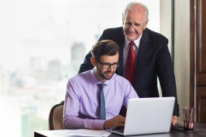 Two businessmen collaborate at a desk: an older man in a suit leans over and smiles at a younger colleague working on a laptop.
