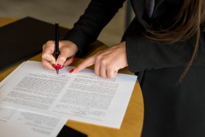 Person signs a document with a black pen on a wooden desk, with a laptop nearby and a second hand resting on the page.