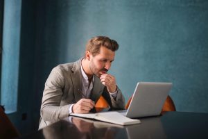 Man in a light blazer sitting at a desk, writing in a notebook beside an open laptop, deep in thought