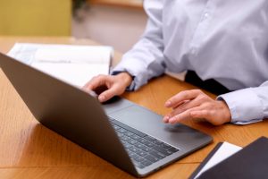 Person typing on a silver laptop at a wooden desk with an open notebook nearby.